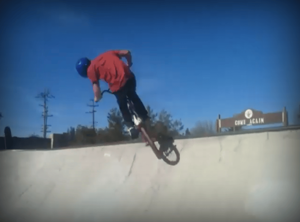 Screenshot of a YouTube video, showing a teenager on a BMX bike, slightly launched into the air off a concrete half-tube in a skate park. His pants are black, shirt is red, and helmet is blue