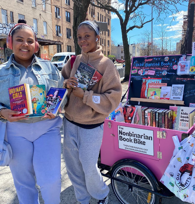 Two Black femmes stand next to the Nonbinarian Book Bike and hold up their book selections at an April pop-up by the Brooklyn Public Library Saratoga branch