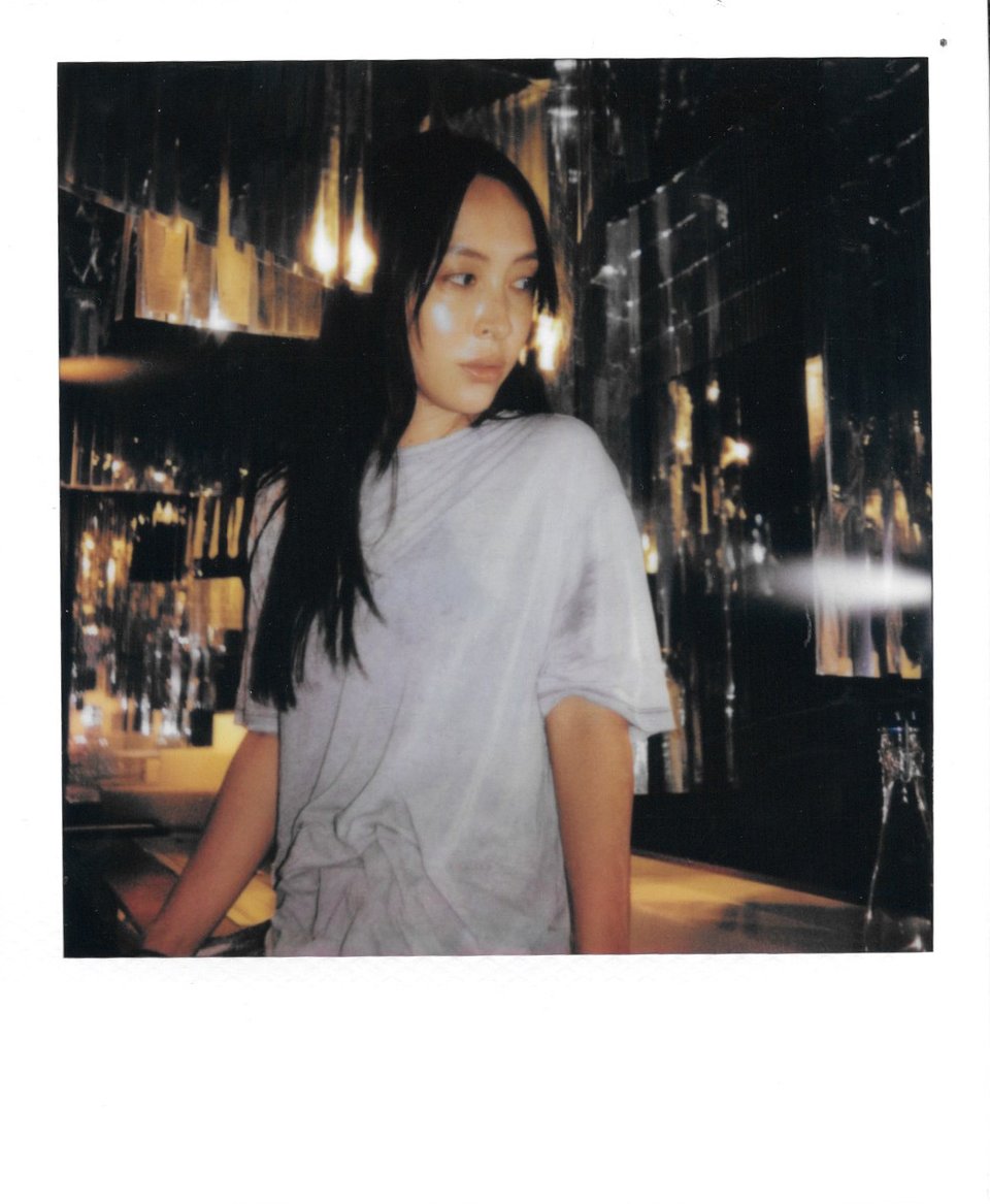 A Polaroid image of artist-filmmaker Diane Severin Nguyen wearing a gray T-shirt and standing in front of a bar or other sparkly/reflective backdrop. Photo is from the waist up, she looks over her shoulder at something out of the frame. Cheekbone has a dramatic highlight on it as though catching reflection of very bright light/effect of Polaroid film
