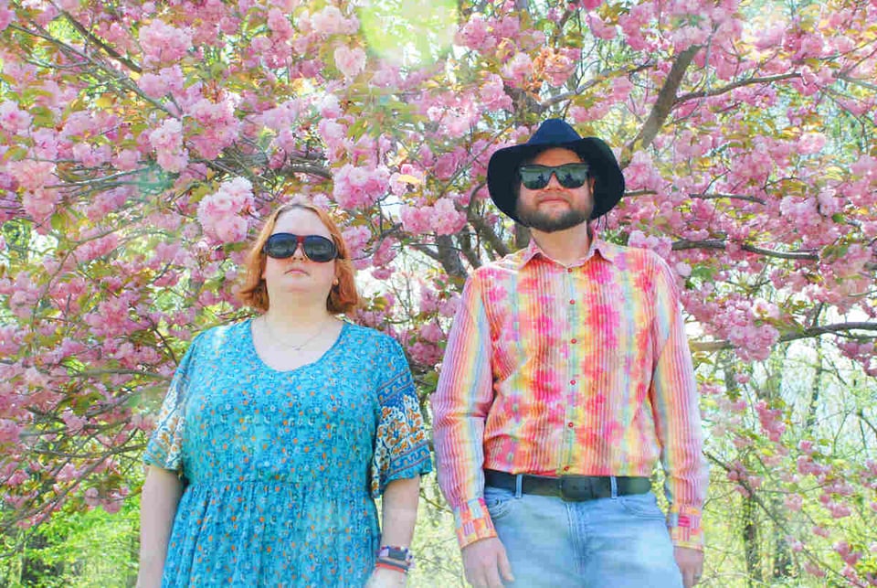 A woman in a blue dress and sunglasses stands next to a man wearing blue jeans, a pastel rainbow-striped shirt, a fedora and sunglasses. They are standing in front of a tree covered in pink flowers.