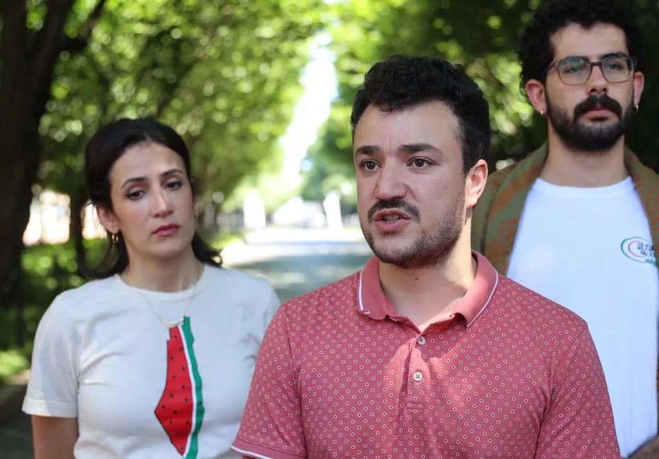 Mahmoud (middle) stands in front of two other individuals while talking at a press conference. They’re outdoors, with trees visible behind them.