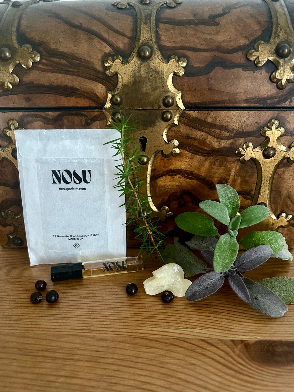 A small spalted walnut treasure chest has a small glass perfume vial with Nosu branding in front of it. There are juniper berries, a sprig of juniper, a nugget of frankincense and some bundles of fresh sage. Image by Rowan Ambrose..