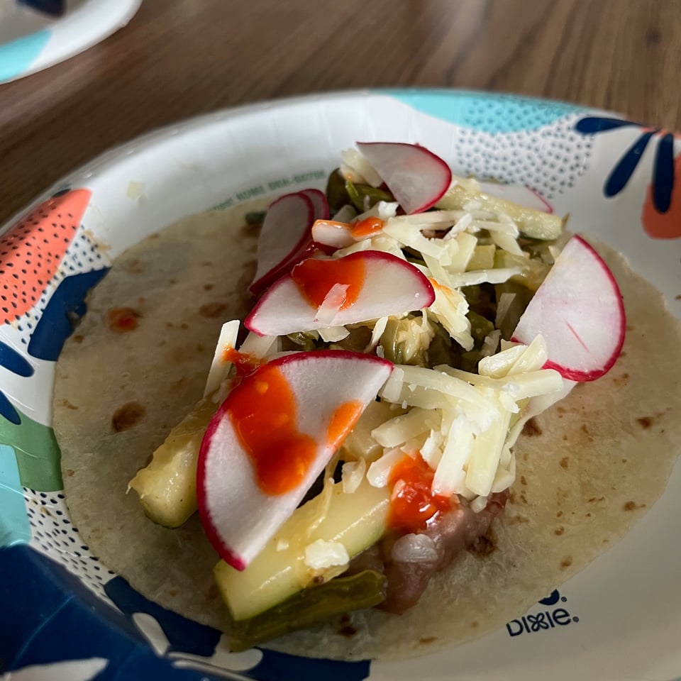 a soft shell taco topped with refried beans, a mix of sautéed nopales and zucchini, cheddar cheese, radishes, and hot sauce