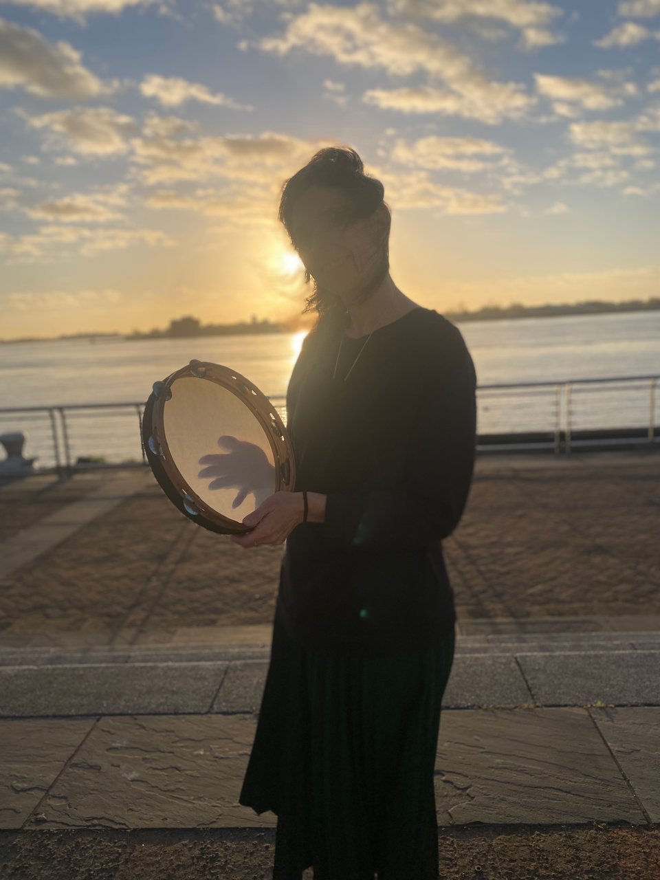 A silhoutted figure holding a large tambourine stands in front of the sun rising over a river.