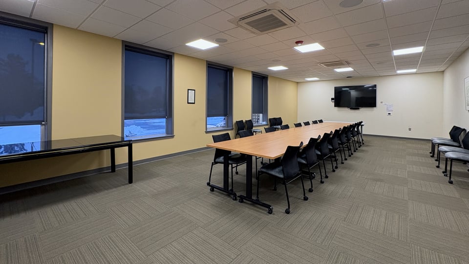 An room with an empty table along a wall at one end and a TV screen mounted on a wall at the other end. A long rectangular table setup has rolling chairs down the length of both sides, with additional chairs against a wall.