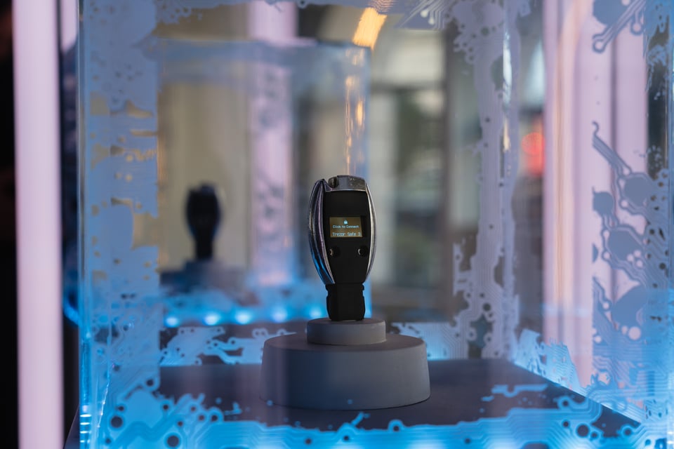 A close up photo of the Key Exchange sculpture inside a glowing display case. It looks just like a smart car key but has a cut out area for a screen that shows the barely visible Tresor logo