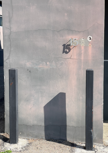 A purplish-gray exterior stucco-ish wall, with only two metal numbers left attached to it from its four-digit address: "15". The sun is shining at a 45º from the upper-right corner of the image, casting the shadow of the "15" onto the wall much more noticeably than the washed-out numbers. The wall is streaked pinkish beneath the area where all four digits were — and streaked more below the two numbers that are still present. There are also two square black metal parking bollards about 3 inches away from the wall, and the one on the right casts a wide black shadow just off-center on the wall. The wide shadow is wedge-shaped and gives an impression of the monolith from 2001: A Space Odyssey