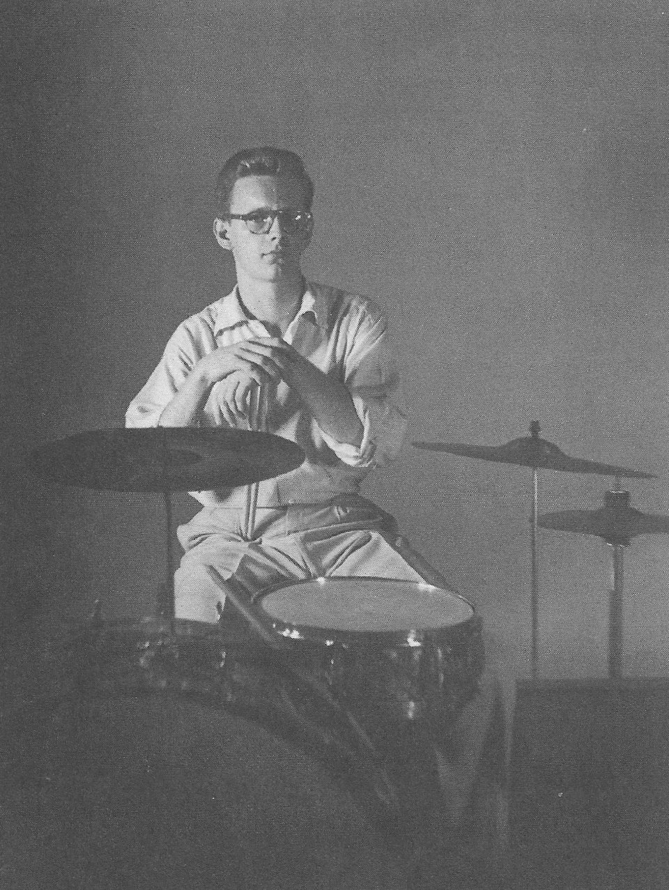 A photograph of a young man, in what appear to be typical 1950s shirt and chinos and eyeglasses, seated behind a minimal drum kit of kick drum, snare, hi-hat and two cymbals. He is holding his drumsticks vertically with the tips pushing down into his leg and his hands resting on top
