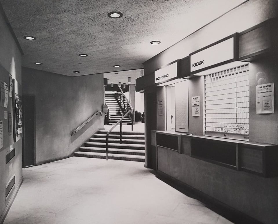 Black and white photo of a theatre foyer. Stairs lead up towards the bar and auditorium whilst a brightly lite box office and kiosk wait for customers. It's all sharp angles.