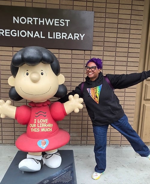 Patricia on one foot with arms spread next to a large statue of Lucy van Pelt, the Peanuts character, who also has her arms spread. There is text on Lucy’s dress that says, “I love our library this much.” They stand in front of the Northwest Santa Rosa Library Branch.
