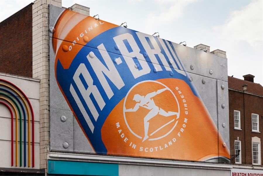Photograph of an IRN-BRU advert posted on a building wall. The ad features a large image of an IRN-BRU can. Under the brand name on the can is the line, “Made in Scotland from girders.” The background of the ad is designed to look like it’s made from steel held together by rivets.