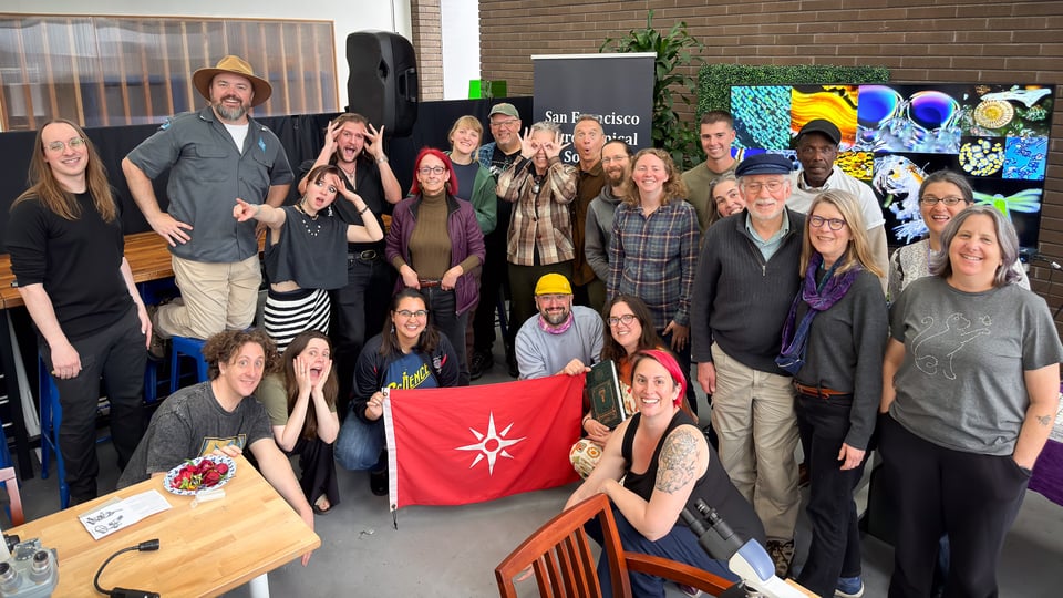 A diverse group of people gathers for a photo, some smiling and making silly faces, with a red flag featuring a star in the foreground.