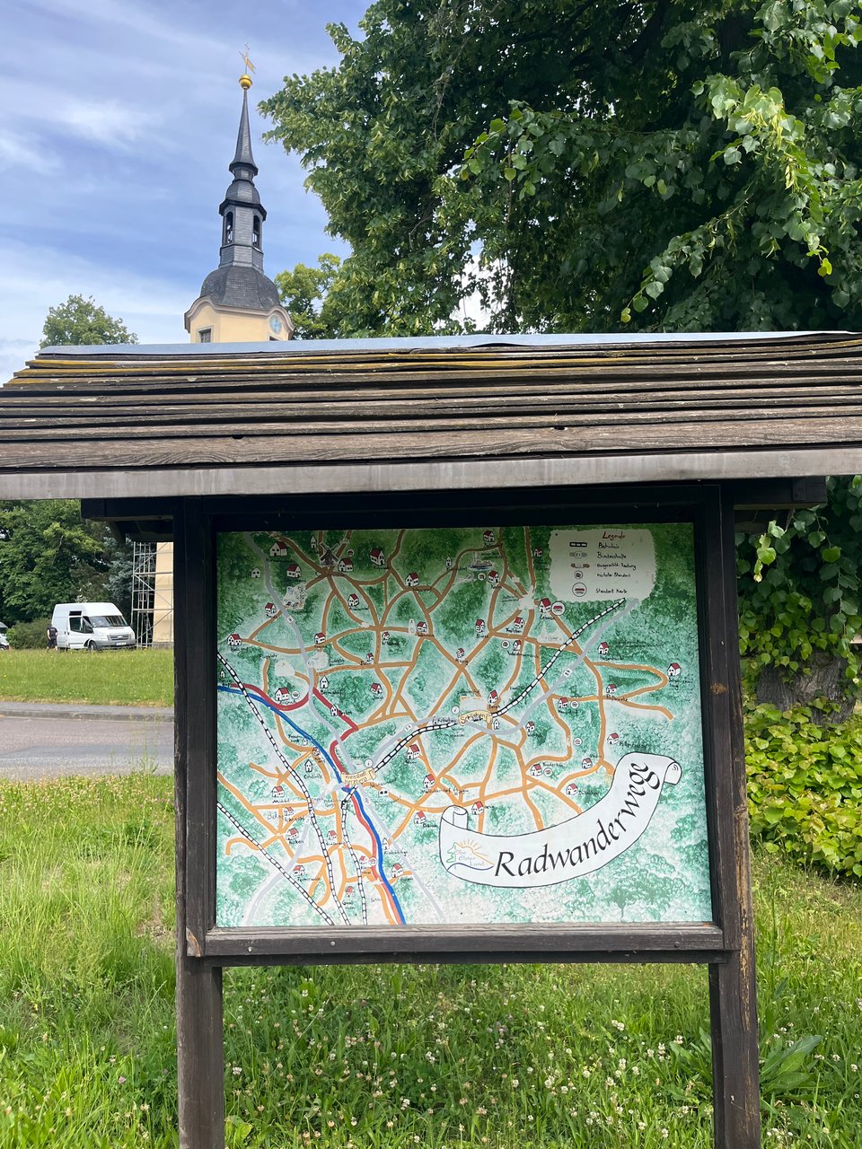 Wooden sheltered sign with hand illustrated map of various paths. Church in background.