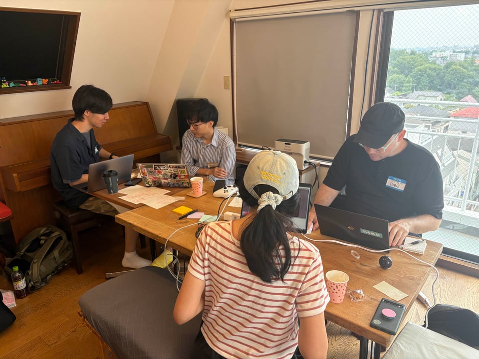 An image of four people gathered around a cluttered workspace. It is bright and sunny outside.