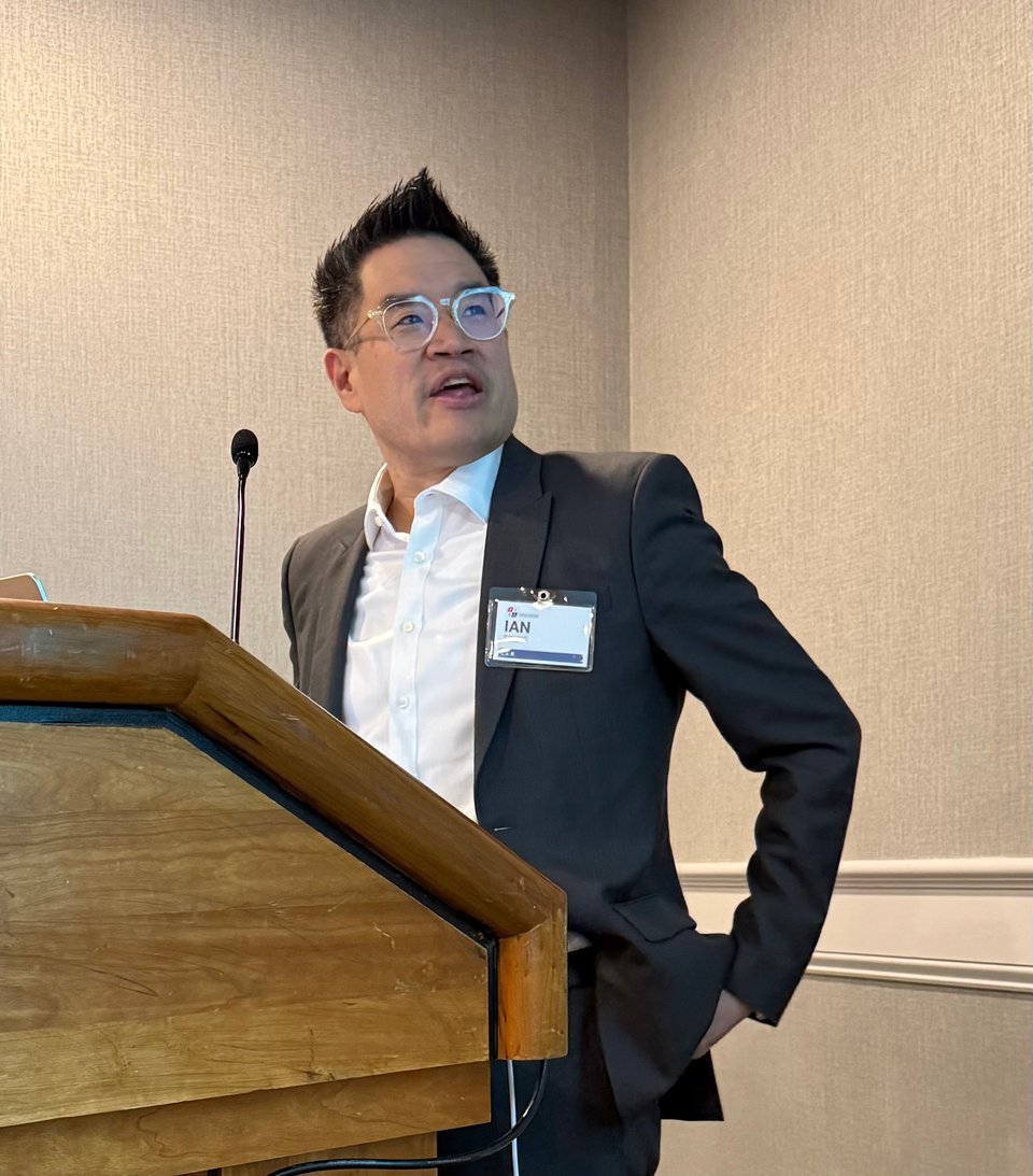 Photo of Asian-American man in gray suit and white shirt at a lectern, turning to look at a projector-screen