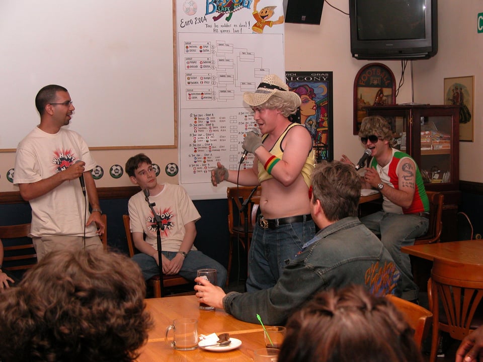 Vinny and Bryan are being interviewed by two characters in outlandish outfits and wigs in a restaurant
