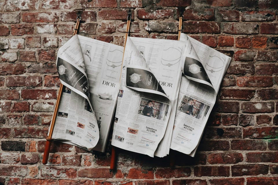 Three newspapers hanging on a brick wall. Photo by Juliana Malta on Unsplash.
