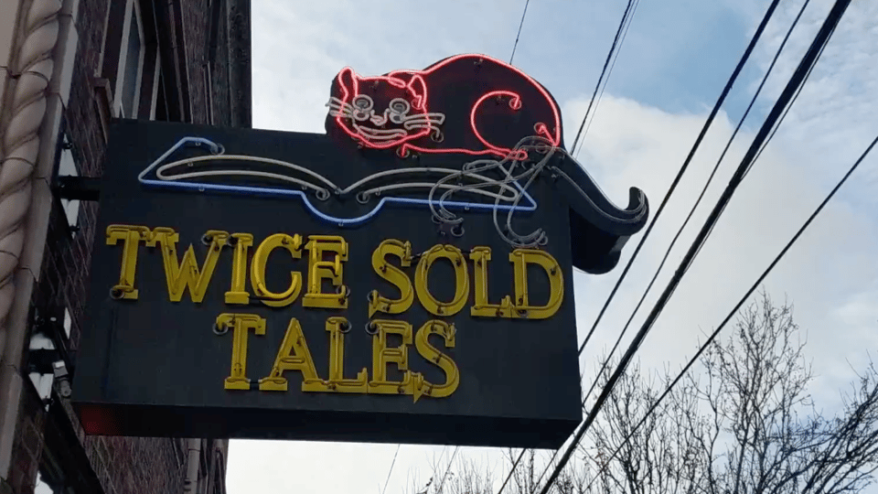A bookstore sign with a grinning pink neon cat looking at an open book. TWICE SOLD TALES is written in yellow gothic script, also in neon.
