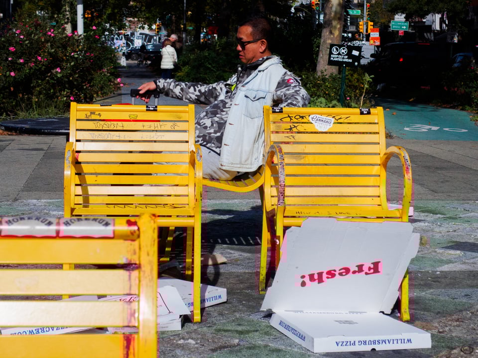 A man sitting and looking cool and chill among yellow public benches. He holds some luggage, and spent Williamsburg Pizza boxes labeled 'fresh!' surround him.