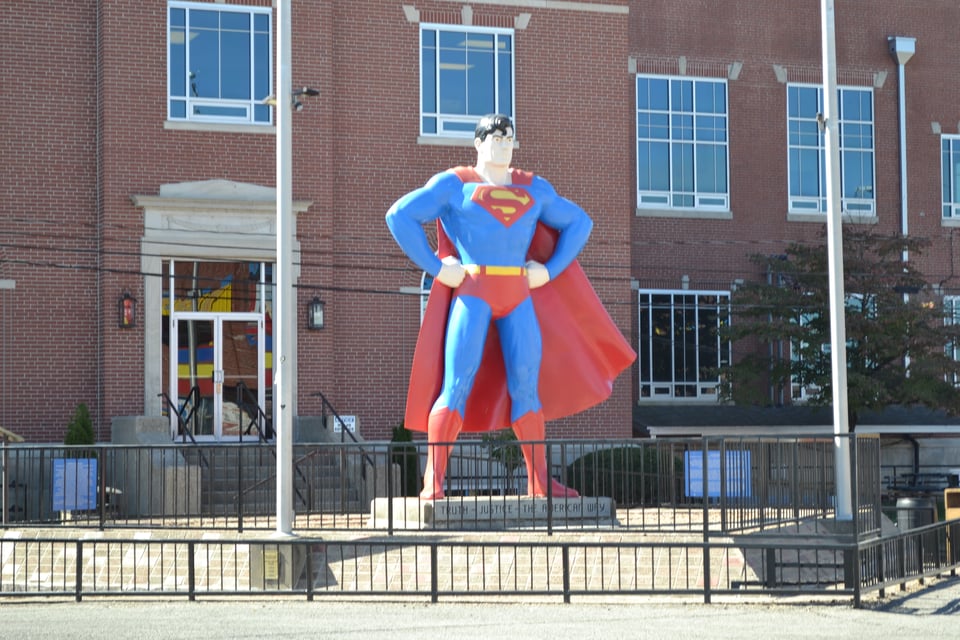 A large statue of Superman, in a classic heroic pose, between two flag plotes and in front of a brick building.