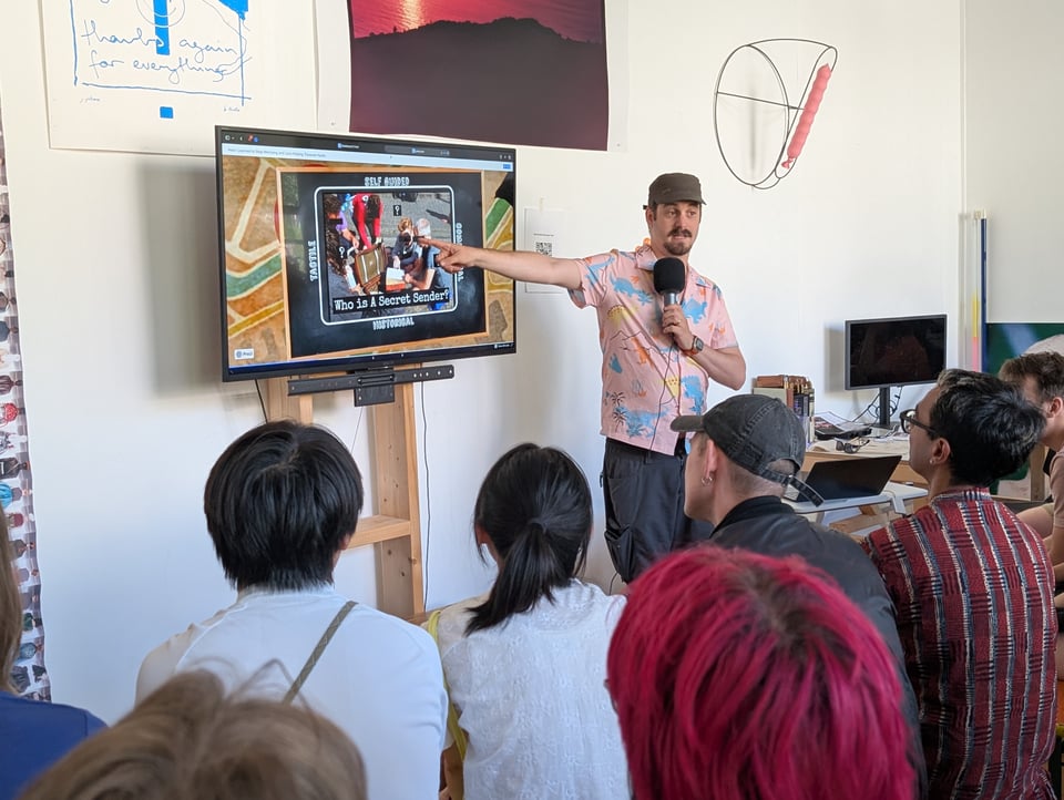A speaker points at a slide on a screen while the audience listens. Text reads "Who is A Secret Sender"?
