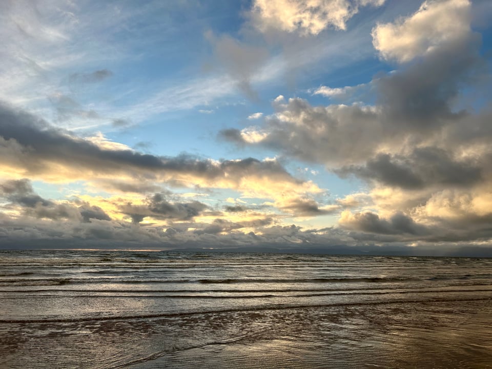 The Isle of Arran and Pladda are in the far distance. The sky is blue with white and grey clouds lit by the setting sun, over a dark grey rippling sea. Image by Rowan Ambrose.