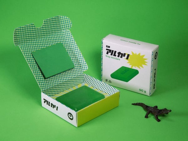 Sobre fondo verde brillante, dos cajas, una abierta y una cerrada. La abierta tiene un patrón geométrico verde y balnco en su interior y una pastilla de jabón verde en su interior. La cerrada tiene una imagen del mismo jabón y carácteres de aspecto asiático. También hay un cocodrilo de plástico.