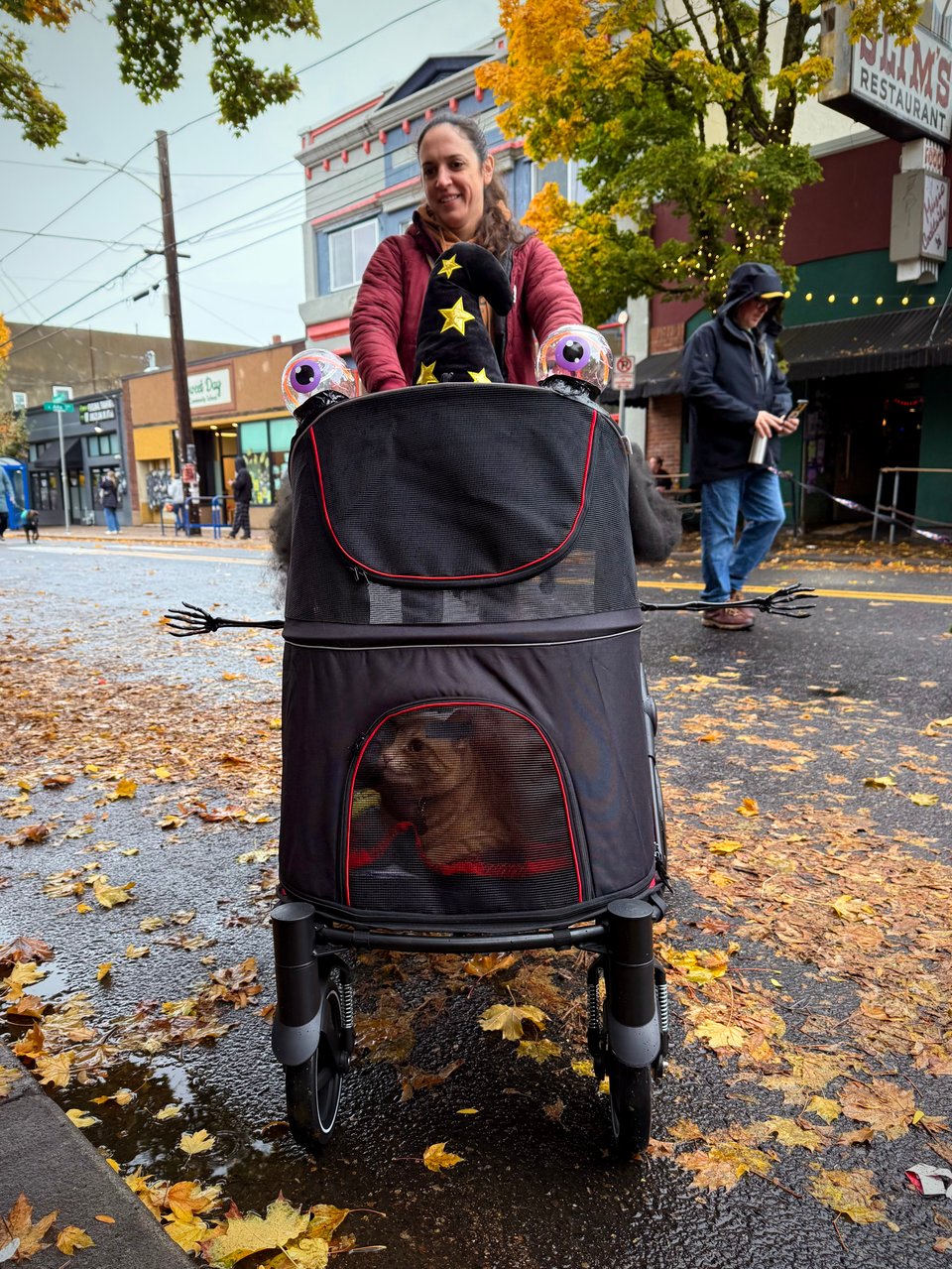 A woman pushes a wheeled carrier with a cat inside. The carrier has eyes, stick skeleton arms, and a wizard hat