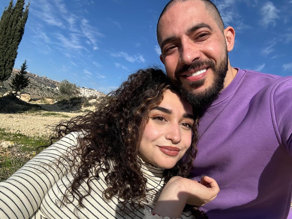 A young woman with long brown curly hair leaning aginast a young man with a beard in a purple shirt; both are smiling