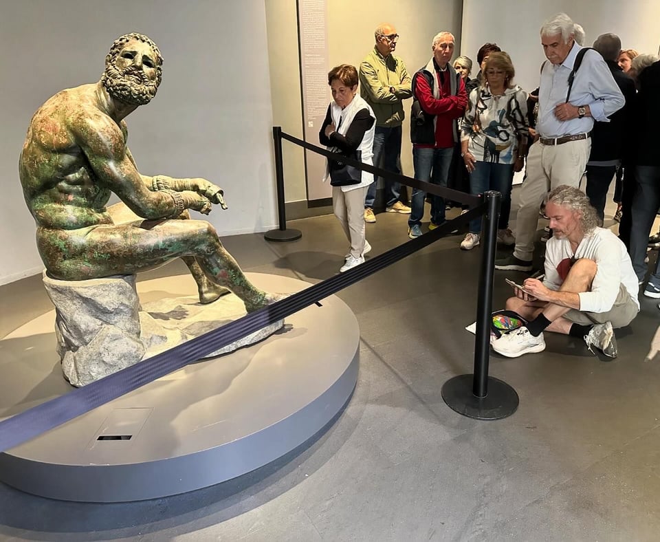 Photo of the statue of The Boxer At Rest; it is blocked off by nylon barrier, and visitors are looking at it. There's some dumb guy sitting on the floor drawing it