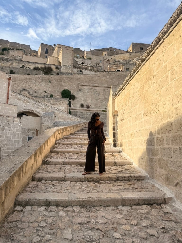 Nadia stands on a wide stone staircase in Matera, dressed in a dark brown outfit, with the historic stone buildings rising behind her in the warm evening light.