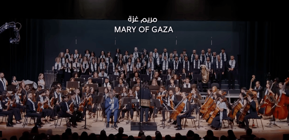 An orchestra and chorus on stage, with the title Mary of Gaza in English and in Arabic