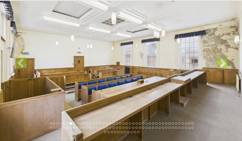 A 1930s courtroom with pale wood benches, docks and justice's bench. Above is a coffered ceiling, and one wall shows signs of damp.