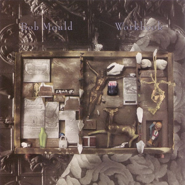 A photograph of a frame with a number of objects placed within. There's too many things to describe, and I'm not sure what all of them are, but I see some shells, a padlock, a figurine of a deer, some clippings, another figurine of a smiling man waving his arm. Behind the frame are some patterns/textures, one looks like it's made of metal and another is a pattern of leaves that looks like it might be from a couch. The whole image is very light grey and light brown.