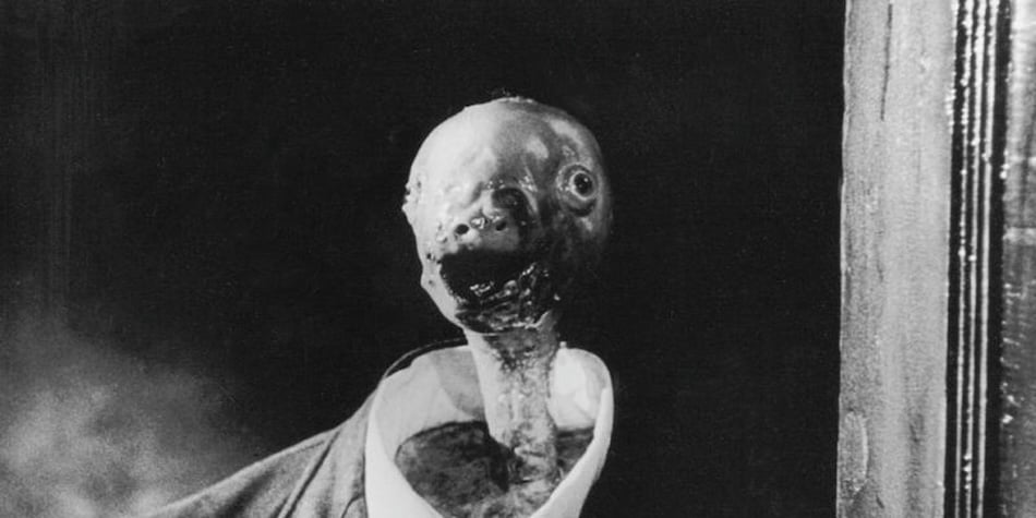A creepy dino baby looking thing with it's head coming out of a giant suit in black and white