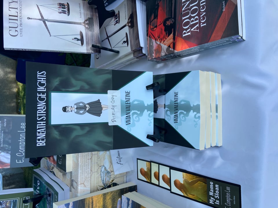 A display of books. The cover depicts a young woman in a gray dress and cardigan standing in a doorway, backlit by bright light. Her shadow transforms into something monstrous. Text reads: Beneath Strange Lights, Vivian Valentine