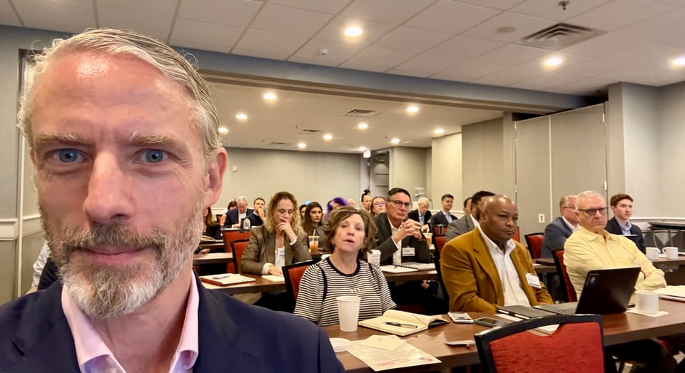 selfie of white man in pink dress-shirt and navy suit, with about 4-dozen people seated behind him in a classroom-style conference room setup