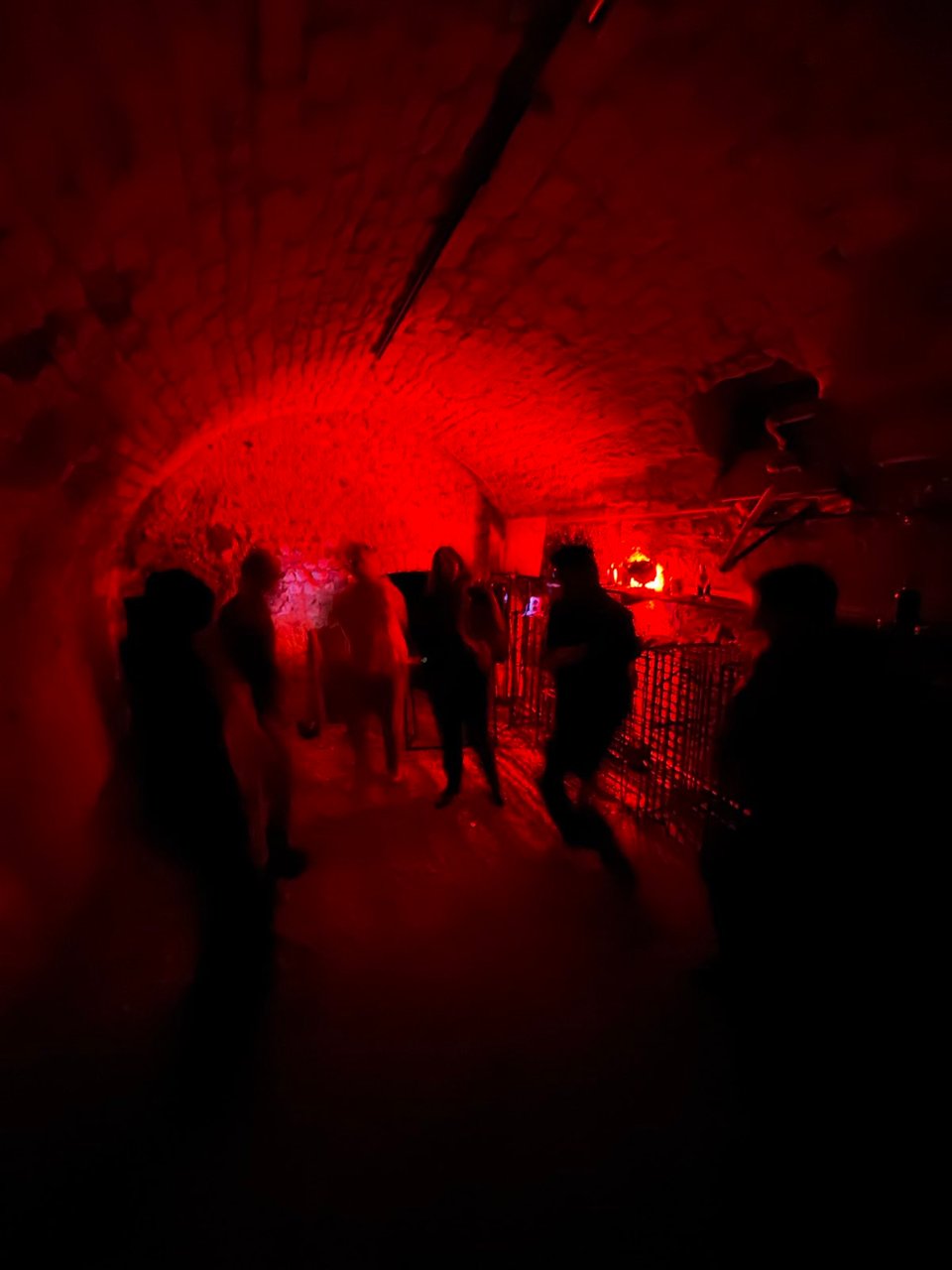 An underground brick cave is lit with red light while six blurry figures dance in the semi darkness.