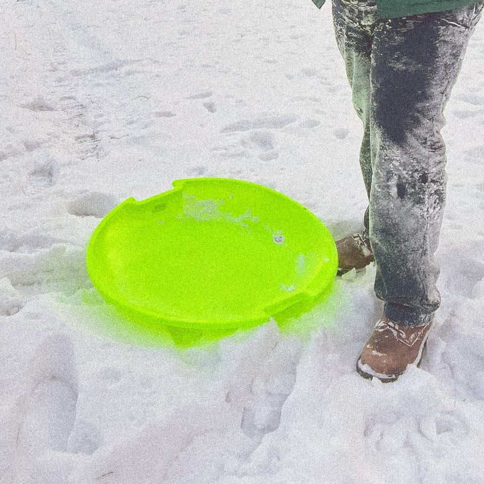 a man in very snowy pants stands next to a tiny neon green sled in the snow