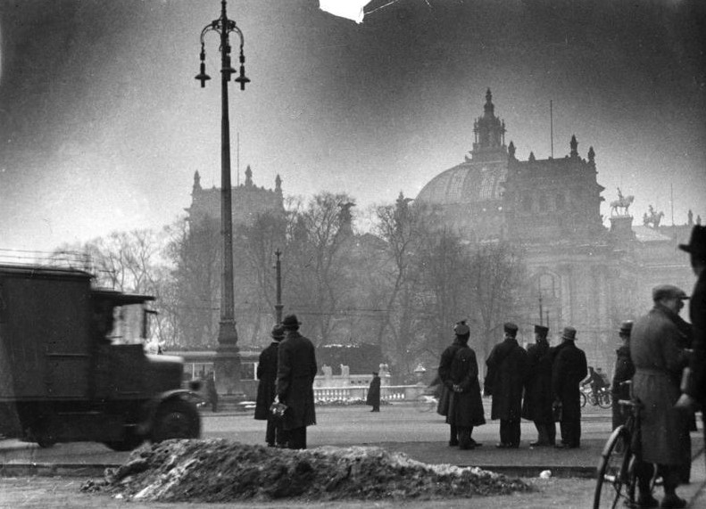A hazy black and white image of a building with a domed roof and cupola, with onlookers silhouetted in the foreground.
