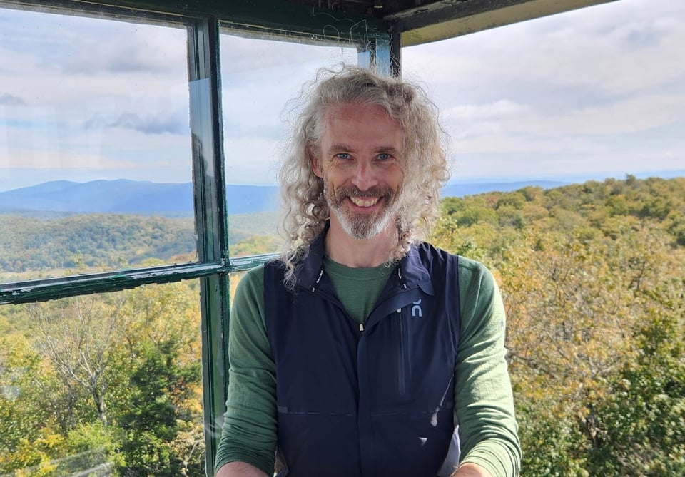 Photo of your humble author in the cab of a fire tower, with trees and mountains in the background. He's smiling, and his long hair is all blown around. He's wearing a green base-layer and a dark running vest