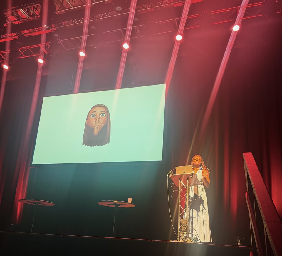 I’m on stage giving a talk, standing behind a podium with my laptop and gesturing as I speak. Red spotlights beam down dramatically, and behind me is a large screen showing my Memoji with a finger to its lips in a “shhh” gesture.