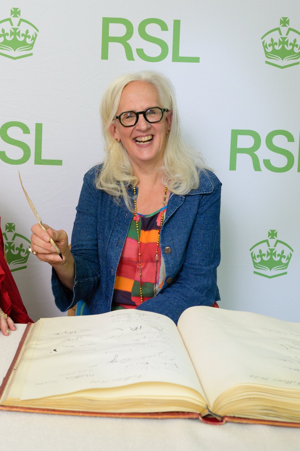 Woman, me, with white hair and glasses in a blue jacket and colourful dress, holding up a quill pen, about to sign my name in the RSL roll book.
