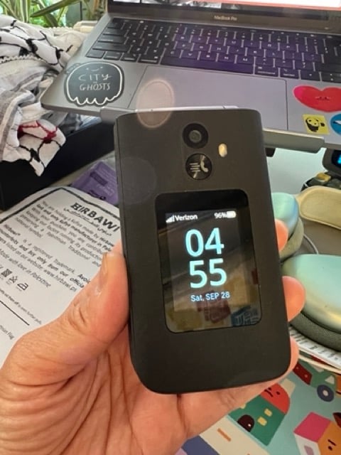 My hand holding a closed flip phone, displaying the time