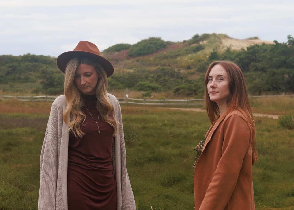 Two women stand in a scrubby natural landscape. The one on the left has blond hair and wears a cowboy hat, a dark earth-tone dress with a lighter sweater. She's looking down. The other, with reddish hair faces the woman on the left, while looking left at the camera. She's wearing an ochre-colored blazer.