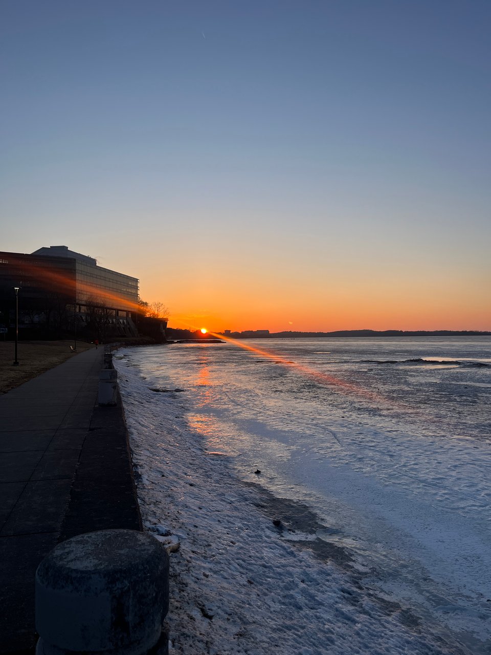 bright orange sunset with diagonal rays of light, orange sky fading into blue, over mostly frozen lakeshore