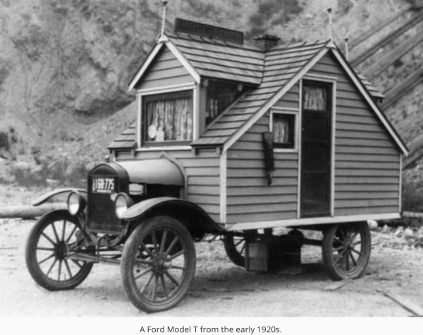 A black-and-white picture of an old fashioned car — kind of like a model T, except where the passenger carriage is supposed to be there is a small house. The overall effect is comical — it looks like a little house that has sprouted wheels and the front engine hood of a motor car.
