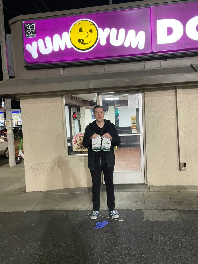 A man in black clutches two bags of donuts outside of a business called Yum-Yum donuts