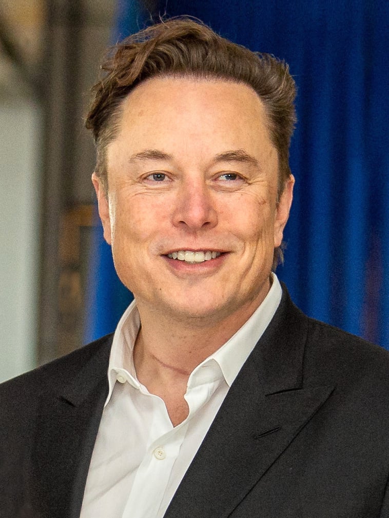 Elon Musk smiles awkwardly while standing in front of a blue curtain. Image via https://www.dvidshub.net/image/7132409/usafa-hosts-elon-musk