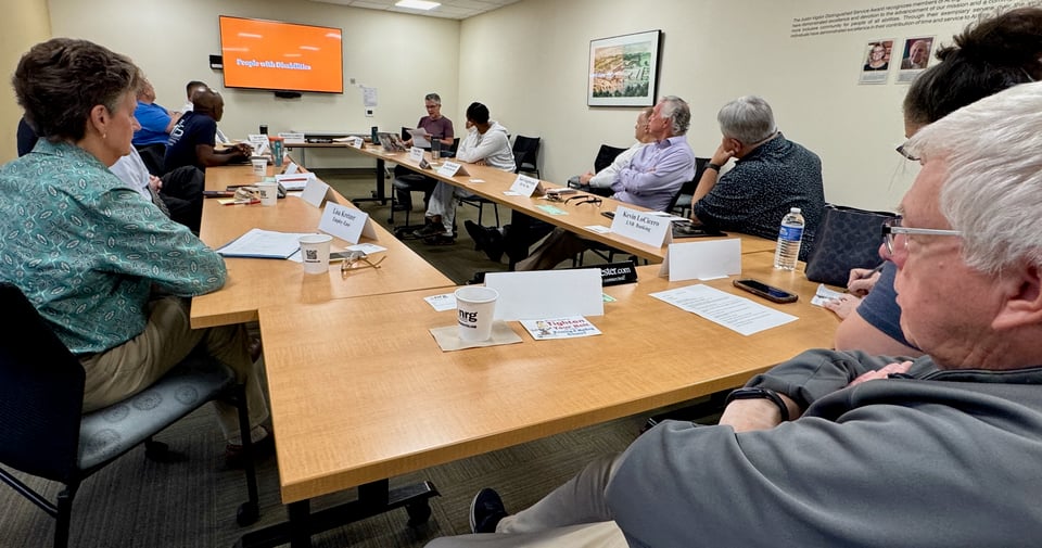 Sara reads her presentation notes to seated NRG members, with an slide that reads People With Disabilities on a wall-mounted display screen.